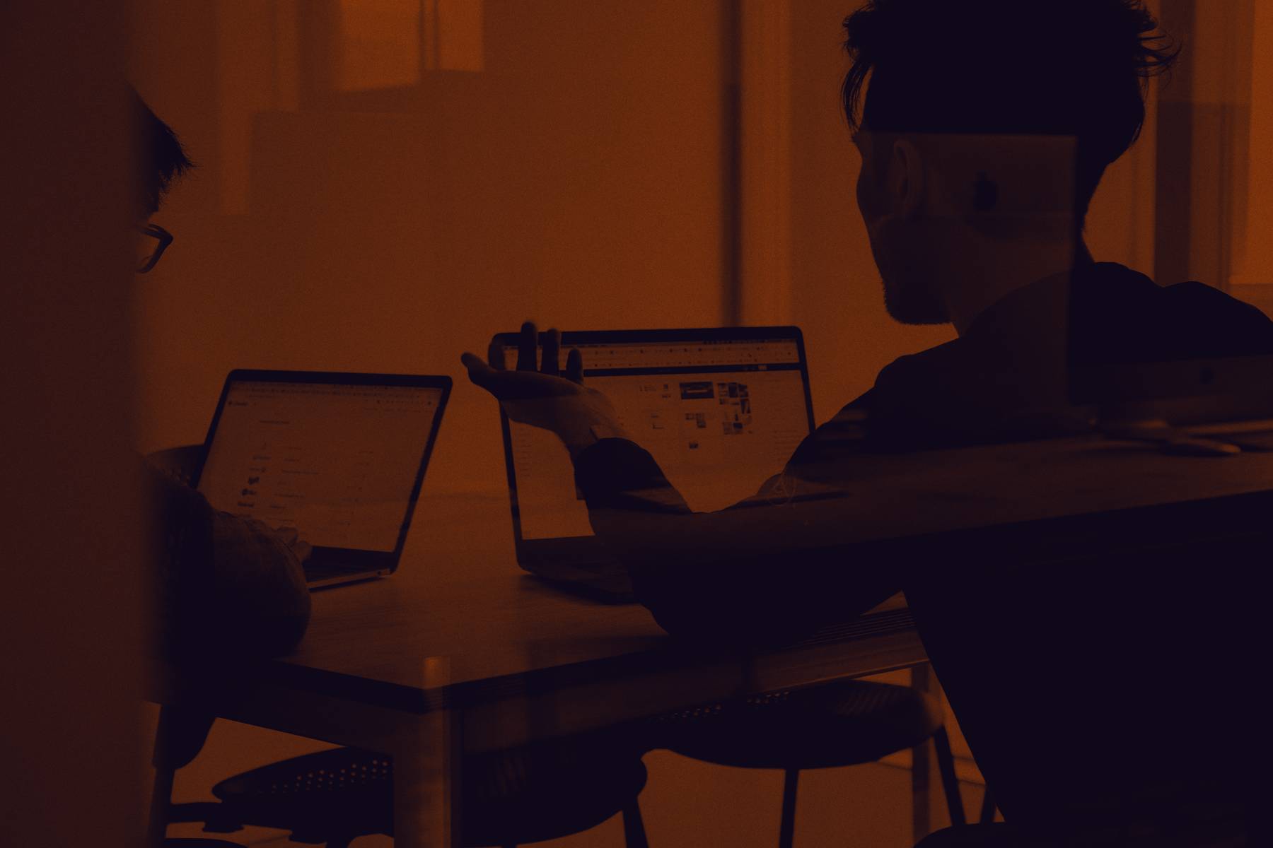 Two people at a table with laptops open between them, photographed through a window